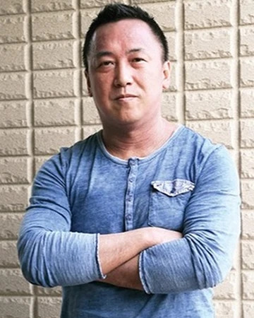 Kuoji Mori standing with his arms crossed, looking into the camera. Behind him is a white wall.